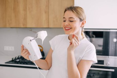 Çağdaş bir mutfak ortamında çırpılmış kremayla elektrikli bir el mikseri tutan neşeli genç bir kadın gülümsüyor.