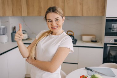 Modern bir mutfakta neşeli bir kadın baş parmağını kaldırır, yemek pişirmekten zevk alır..