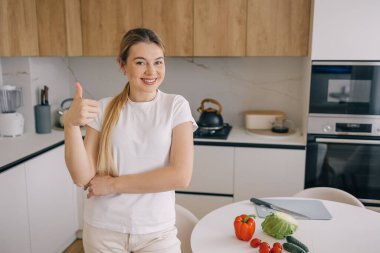 Modern bir mutfakta, masada taze sebzelerle baş parmağını kaldıran, sağlıklı beslenme ve yemek hazırlığında pozitifliği teşvik eden bir kadın..