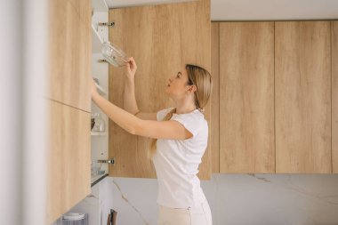 Beyaz tişört giyen bir kadın, düzenli ve düzenli bir yaşam tarzının somutlaştırıldığı, parlak ahşap bir mutfak dolabına eşyalar yerleştiriyor. Parlak ve minimalist mutfak.