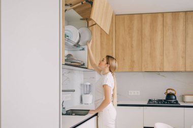 Bir kadın, modern bir ev ortamında mutfak dolabına tabak koyuyor..