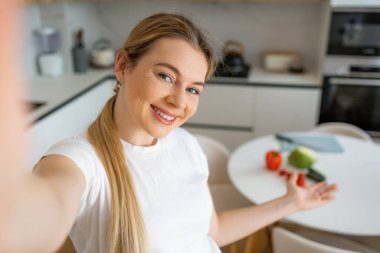 Neşeli bir kadın gülümsüyor ve sıcak bir ev mutfağında sebze gösteriyor. Kendini selfie ile filme çekiyor.