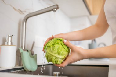 Modern bir mutfakta akan suyun altında yeşil bir marulu yıkayan bir çift el. Sağlıklı beslenme alışkanlıkları ve ev ortamında yemek hazırlama.