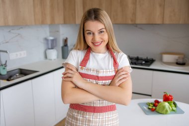 Önlük takan neşeli bir kadın, modern bir mutfakta taze sebzelerle duruyor. Yemek hazırlama ve sağlıklı beslenme ve yaşam tarzına odaklanmayı öneriyor..