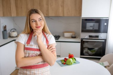Modern bir mutfakta düşünceli bir kadın, taze sebzelerle çevrili, büyüleyici bir önlük giymiş, sağlıklı bir yemek planlıyor..