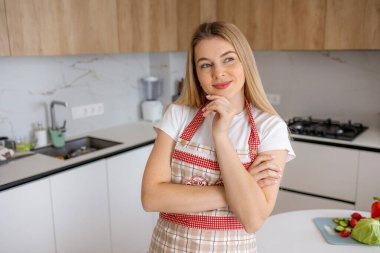 Önlük giymiş genç bir kadın, mutfakta dikiliyor, düşünceli ve ilham verici görünüyor.