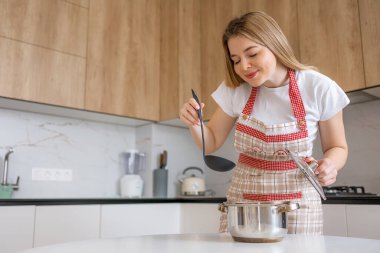 Önlük takan neşeli bir kadın şık bir mutfakta tencerede yemek karıştırıyor..