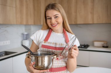 Önlüklü gülümseyen kadın, yemek pişirme ve hazırlama zevkini temsil eden, parlak, modern bir mutfakta bir kap ve kaşık tutuyor..