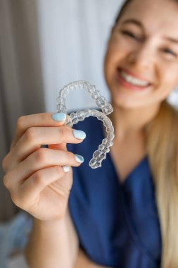Gülümseyen bir birey bir çift şeffaf hizalayıcı sergiler, diş hizalaması için modern ortodontik tedavileri vurgulayarak, oral sağlığı ve estetik olarak tatmin edici