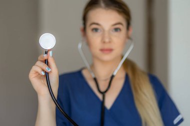 Mavi üniformalı odaklanmış bir tıp uzmanı kameraya stetoskop doğrultuyor. Çeşitli ortamlarda sağlık hizmetlerinin, tıbbi mesleklerin veya klinik uygulamaların resmedilmesi için ideal.