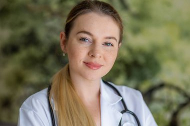 Beyaz önlük ve steteskop giyen, özgüven ve sıcaklığı ifade eden, doğal yeşil arka planda sakin ve dostane bir atmosfer yaratan genç bir kadın sağlık uzmanının portresi..