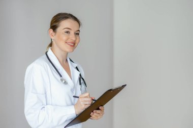 Profesyonel bir kadın doktor, panoya gülümseyip steteskop ile beyaz önlük giyip, sağlık hizmetleri ortamında tarafsız bir arkaplanda duruyor.