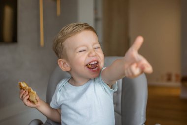 Neşeli genç bir çocuk evde atıştırmalık yiyor, bir sandalyede rahatça otururken neşe ve neşe ifade ediyor, içten bir kahkaha ve kaygısızlıkla mutluluğu yayıyor..