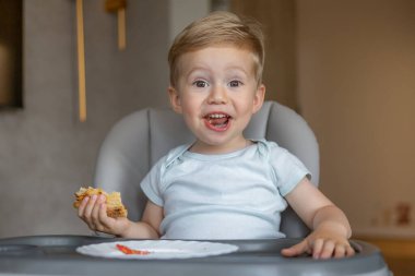 Neşeli genç bir çocuk sandalyeye oturmuş, elinde yemek tutuyor ve mutlu bir yüz ifadesi sergiliyor, yemek zamanını yakalıyor.