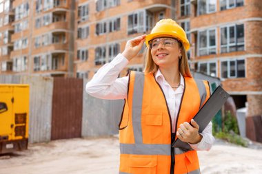 Kasklı ve yansıtıcı yelek giyen bir kadın inşaat alanında bir pano tutuyor. konut inşaatı, mühendislik ve inşaatta ilerleme, güvenlik protokollerinin korunması. 