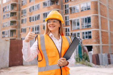 Yansıtıcı giysi ve başlık takan kendine güvenen bir kadın inşaat işçisi aktif bir inşaat ortamında başparmağını kaldırıyor.. 