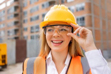 Kasketi ve güvenlik yeleği olan kadın inşaat işçisi inşaat alanındaki gelişmekte olan binaların arasında güvenle gülümsüyor. 