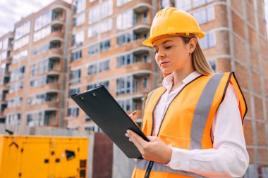 İnşaat sahasında, arka planda inşaat halindeki bir bina ile birlikte inşaat halinde olan ve elinde bir pano tutan sert bir şapka ve yansıtıcı yelek giyen bir kadın işçinin olduğu bir yer.. 