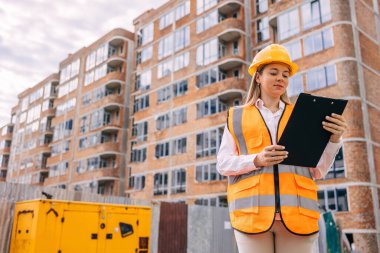 Güvenlik teçhizatı takan bir inşaat işçisi, şantiyede duruyor ve bir binanın yakınındaki belgeleri inceliyor.. 