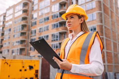 Sert şapka ve güvenlik yeleği giyen bir kadın mühendis, inşaat halindeki modern bir binanın önündeki ilerlemeyi gözlemlemek için, bir pano ile inşaat alanını inceliyor.. 