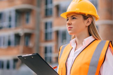 Sarı kasketli ve yansıtıcı yelek giyen bir kadın, inşaat alanını izleyen bir pano tutuyor.. 