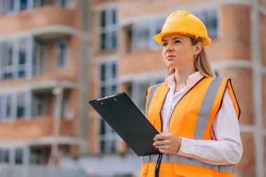 Sert şapka takan ve yansıtıcı yelek giyen bir kadın mimar, inşaat çalışmalarını değerlendirirken elinde bir pano tutan ve uyumu sağlayan bir inşaat projesini gözlemliyor.. 