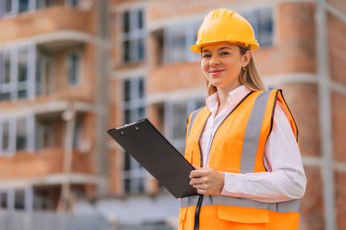 Sarı kask ve güvenlik yeleği giyen, elinde siyah bir dosya tutan gülümseyen bir kadın inşaat yöneticisi. Kentsel gelişimde ilerleme ve sanayi profesyonelliği. 