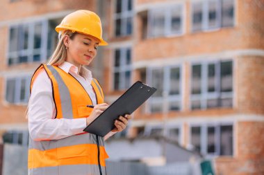 Güvenlik kaskı ve yelek giyen bir kadın inşaat işçisi dışarıda teftiş görevleri yapıyor. Elinde bir pano tutuyor, bir inşaat alanının yanında durup gözlemleri belgeliyor.. 