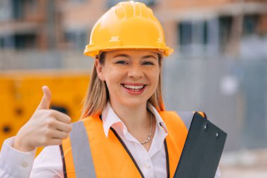 Sıkı bir şapka ve güvenlik yeleği giyen neşeli bir inşaat işçisi inşaat alanında dikilirken başparmak hareketi yapıyor. 