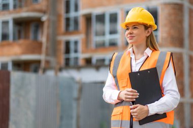 Profesyonel inşaat kıyafeti giymiş bir kadın elinde bir panoyla şantiyede duruyor.. 