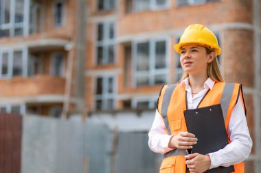 Profesyonel inşaat kıyafeti giymiş bir kadın elinde bir panoyla şantiyede duruyor. 