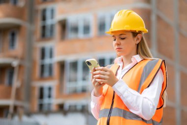 Başlık ve güvenlik yeleği giyen bir inşaat işçisi, inşaat sahasının dışında akıllı telefon kullanıyor ve elindeki mobil cihaza odaklanıyor.. 