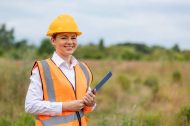Kendine güveni tam bir inşaat işçisi, turuncu bir güvenlik yeleği ve bir tarlada duran sert bir şapkası var.. 