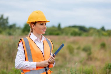 Açık hava şantiyesinde duran bir kadın sarı bir şapka ve turuncu bir güvenlik yeleği giyiyor. İlerlemeyi ya da planları incelemek için elinde bir pano tutuyor.. 