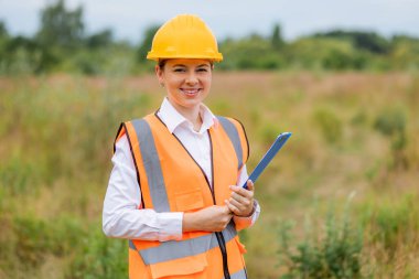 Kasketli ve yansıtıcı yelek giyen bir kadın işçi kırsal kesimde bir pano tutuyor.. 
