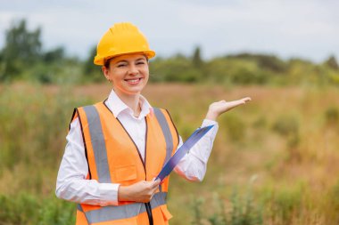Koruyucu teçhizatlı, elinde pano olan, açık hava inşaat alanına doğru hareket eden kendine güvenen bir kadın.. 