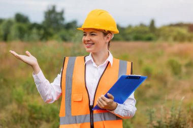 Kendine güvenen kadın mühendis güvenlik ekipmanı giyip dışarıda elinde bir pano tutuyor. Profesyonellik, güvenlik yönetmeliklerine uyum ve açık hava iş denetimleri sırasında inşaat veya mühendislik alanında pozitiflik. 