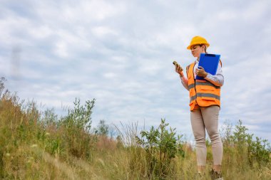 Yansıtıcı yelek ve kasketli bir işçi açık havada telefon ve pano kullanıyor. 