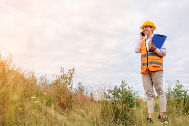 İnşaat işçisi, yüksek görüş açısına sahip yeleğiyle, elinde pano, cep telefonuyla konuşuyor.. 