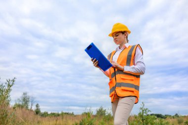 Sert şapka ve yansıtıcı yelek giyen bir kadın açık hava ortamında belgeleri inceliyor. Arka plandaki bulutlu gökyüzü ve çimenli alan inşaat veya açık hava projelerinin çalışma ortamını vurgular. 