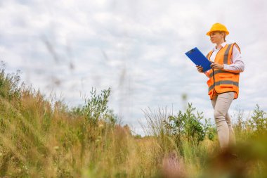 Güvenlik yeleği ve sarı kask giyen bir kadın mühendis, bir pano tutarken cep telefonuyla iletişim kuruyor, çimenli bir alanda dışarıda duruyor. 