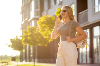 Güler yüzlü bir kadın güneşli bir şehirde telefonunu kullanarak dışarı çıkıyor. Akıllı telefonuyla konuşuyor, günbatımında şık bir çantayla sıradan giyiniyor.
