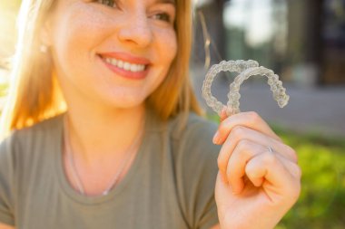 Mutlu genç bir kadın güneşli bir park ortamında temiz diş bakımı, ortodonti ve modern diş hekimliği gösteriyor..