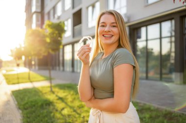 Neşeli bir kadın gündelik kıyafetlerle açık bir hiza tutuyor, açık şehir ortamında canlı güneş ışığı ve yeşillikle gülümsüyor, modern ortodontik çözümleri ve olumlu duyguları resmediyor..