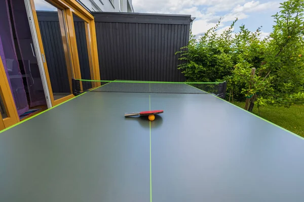 View of tennis ping pong table with net and racket with orange ball in yard of private house on summer day. Sweden.