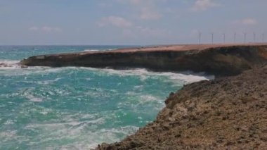 Gorgeous view of rocky coast, where big turquoise waves of Atlantic ocean crash against rocks against backdrop of wind turbines in desert. Aruba.