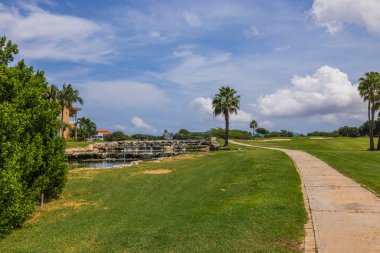 Güzel tropikal doğa manzarası. Mavi gökyüzünde gölet ve golf sahası arasında beyaz bulutlu bir yol var. Aruba. 