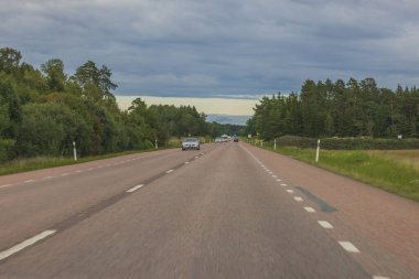 İki tarafında yeşil ağaçlar olan asfalt yol mavi bulutlu gökyüzü ile birleşiyor. İsveç.