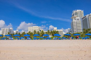 Mavi güneşli ve şemsiyeli Miami 'nin kumlu sahili beyaz binalara karşı, hepsi mavi gökyüzünün altında, yumuşak bulutlu. Miami Plajı. ABD. 