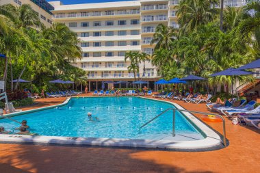 Misafirler sıcak yaz gününde, yeşil palmiye ağaçlarıyla çevrili, merkez otel havuzunun yanındaki parlak mavi şemsiyelerin altında güneşleniyorlar. Miami Beach, Florida, ABD. 